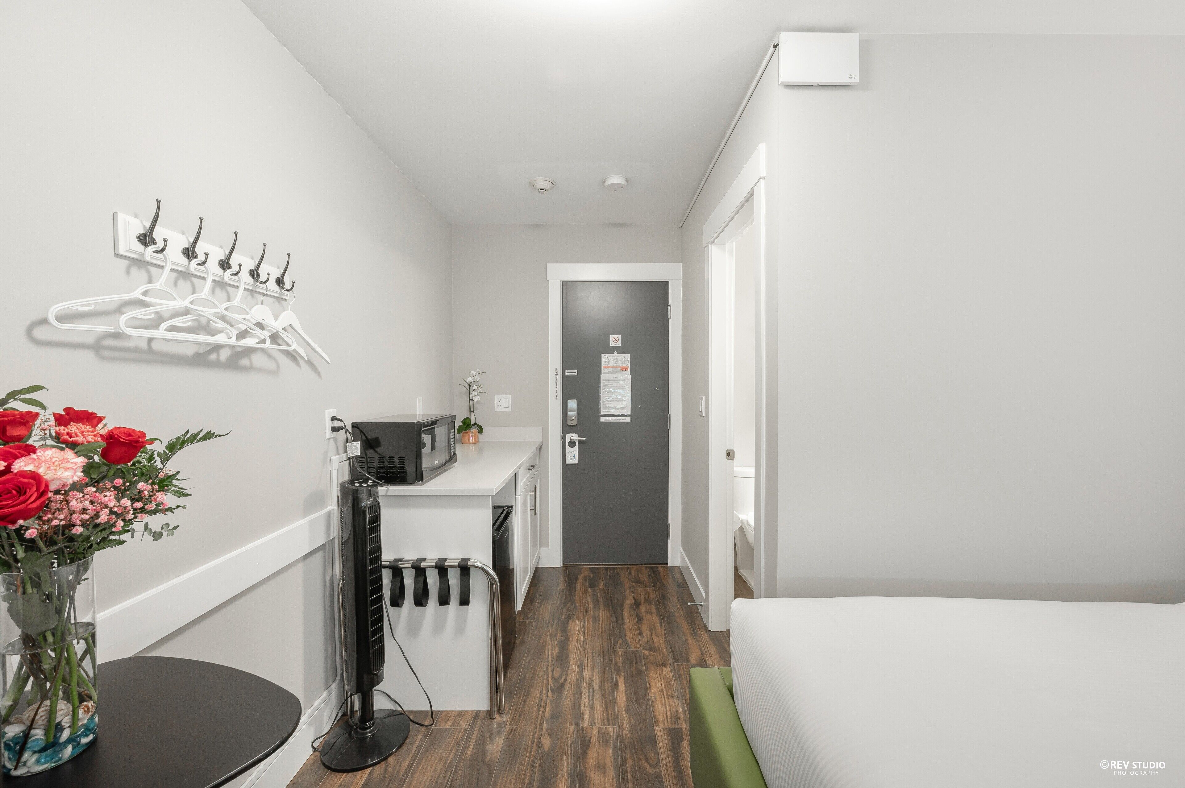 A modern hotel room with a bed, a small kitchenette, a microwave, a coffee machine, a black fan, and a door with a sign.