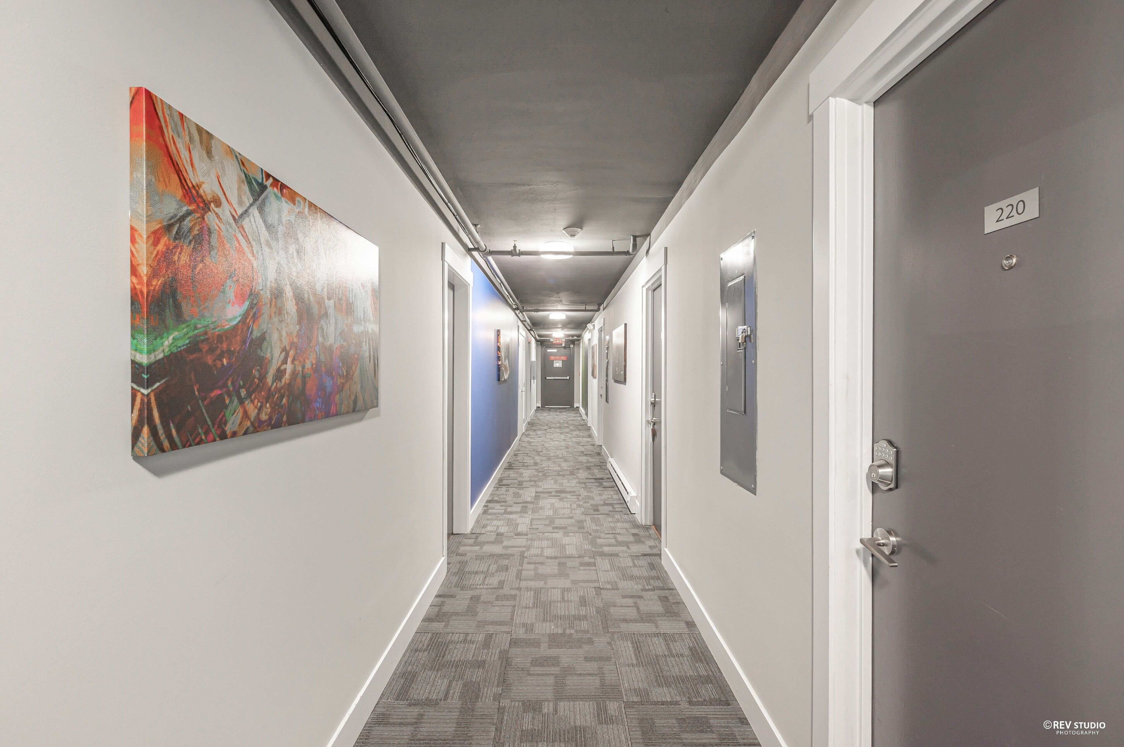 Pasillo de hotel con alfombra estampada, un cuadro abstracto grande en la pared izquierda y puertas numeradas 220.