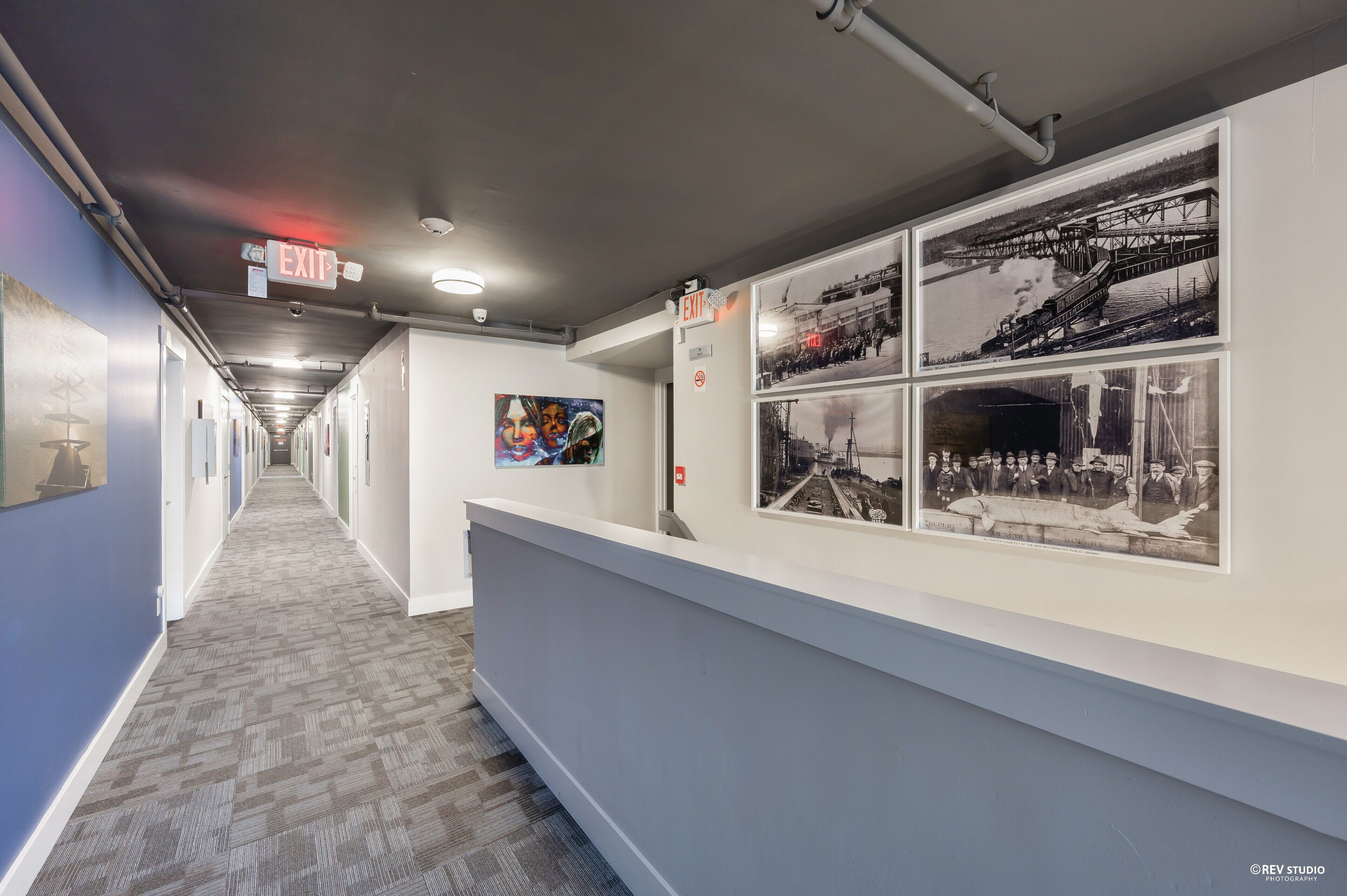 A hallway with an exit sign, a row of framed photographs on the wall, and a patterned carpet.