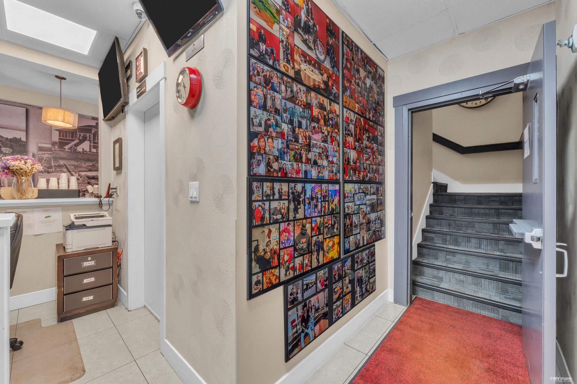 Un pasillo con un amplio collage de fotos en la pared, una escalera con alfombra roja y un escritorio con una impresora.