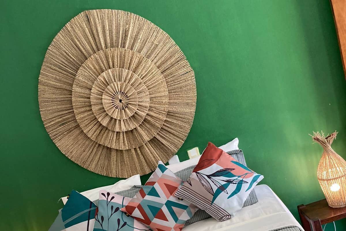 A neatly made bed with a green bedspread, decorative pillows, a woven wall hanging, and a small table with a lamp.