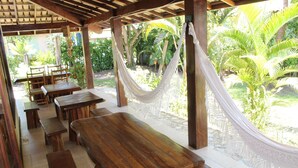 A covered outdoor dining area with wooden tables and benches, surrounded by lush greenery and a hammock. - Pousada Velas e Vento - Acesso fácil a Barra Grande (Maraú)