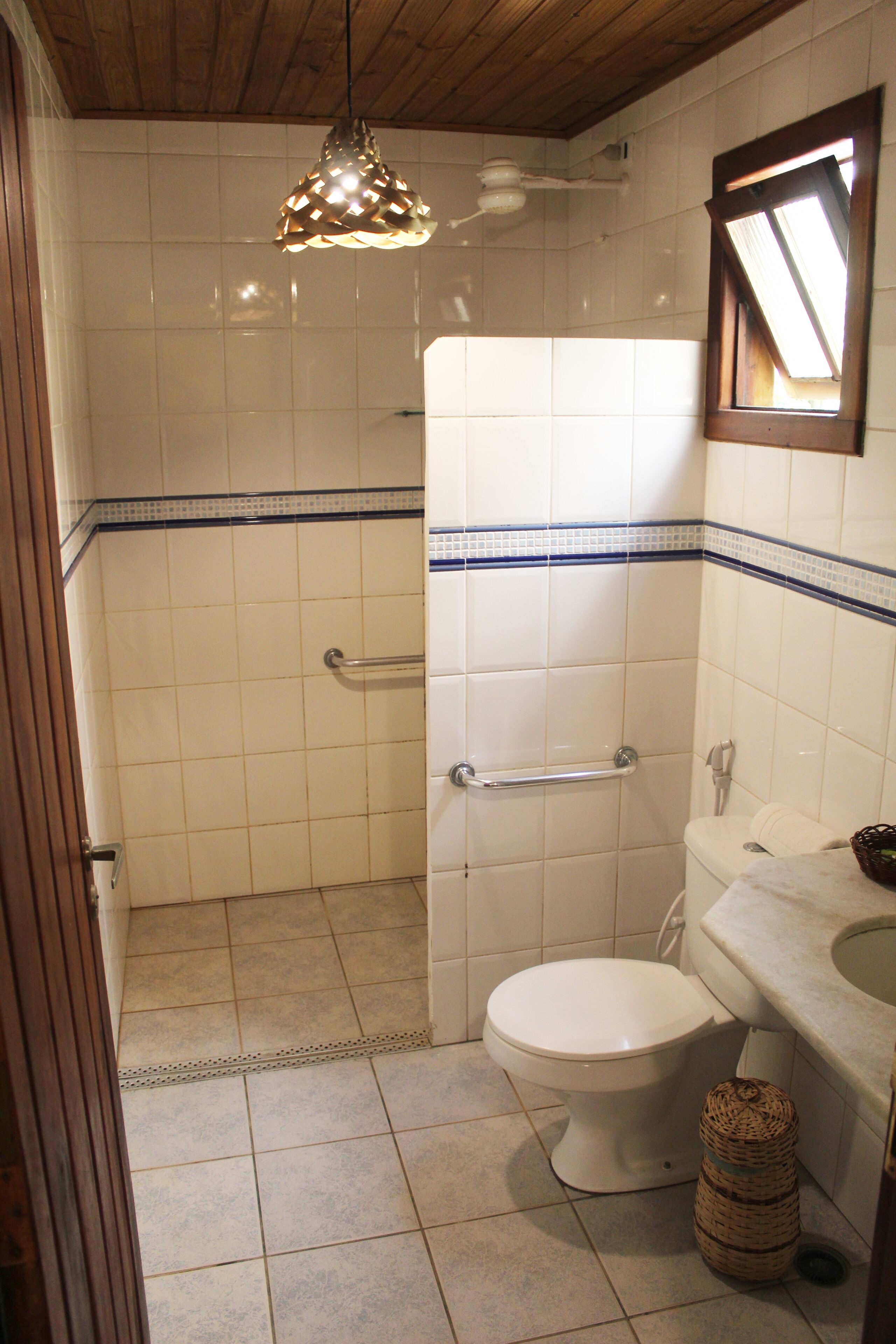 Un baño con inodoro blanco, ducha con barra de apoyo, techo de madera y lámpara colgante.