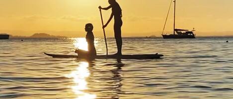 A sunset over calm water with a person standing on a paddleboard and another on a boat.