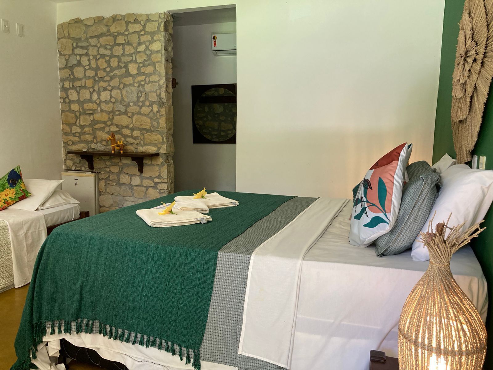 A hotel room with a stone wall, a bed with a green blanket, multiple pillows, and a decorative vase.