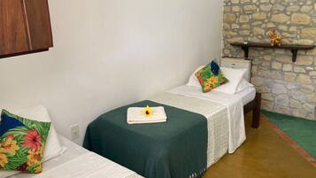 A room with two single beds, each with a green bedspread and a floral pillow. There is a small shelf with decorative items on the wall.