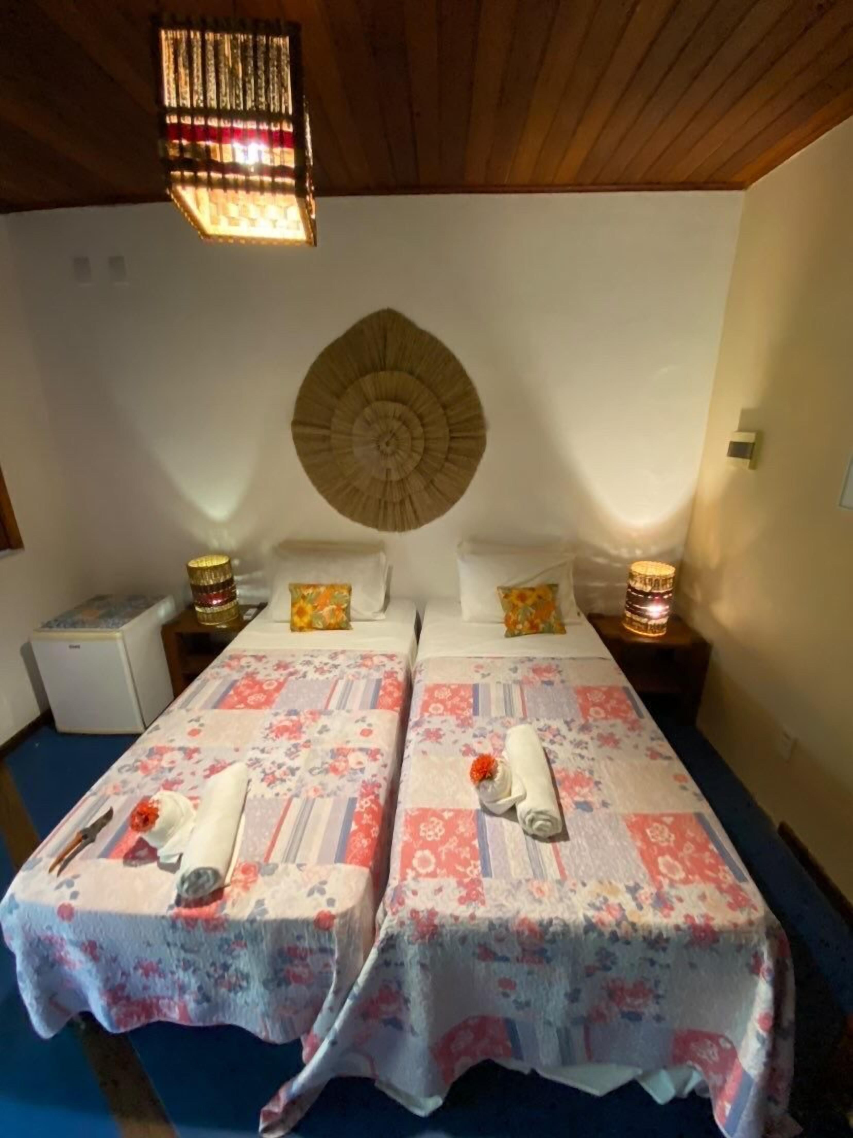 Une chambre double avec deux lits, chacun agrémenté de draps à motifs floraux et d’une serviette roulée. Un élément décoratif orne le mur au-dessus des lits, et une petite table avec une lampe se trouve entre les deux.