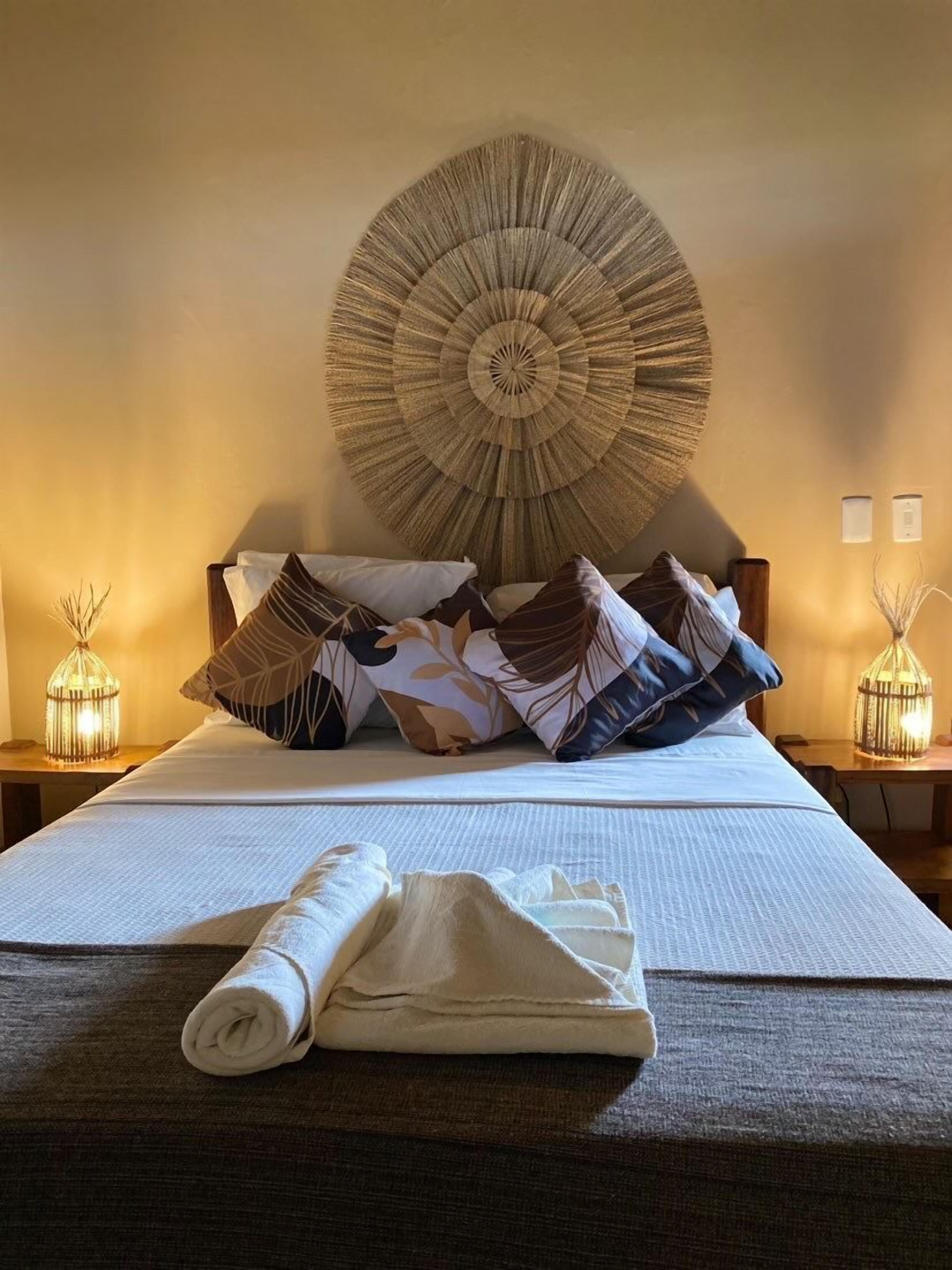 A neatly made bed with a large, circular wall hanging above it, two bedside tables with lamps, and a rolled towel on the bed.