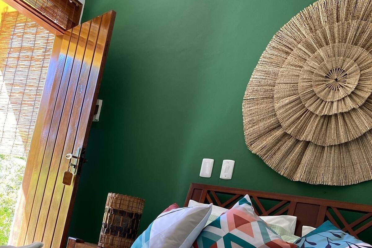 A neatly made bed with a rolled white towel, a green wall, a wooden door, and a woven wall hanging.