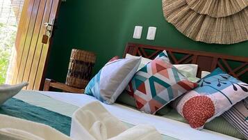 A neatly made bed with a rolled white towel, a green wall, a wooden door, and a woven wall hanging.