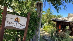 A wooden sign with the text "Velas e Vento Pousada" in a rustic outdoor setting with a stone pathway, greenery, and a tiled-roof structure. - Pousada Velas e Vento - Acesso fácil a Barra Grande (Maraú)