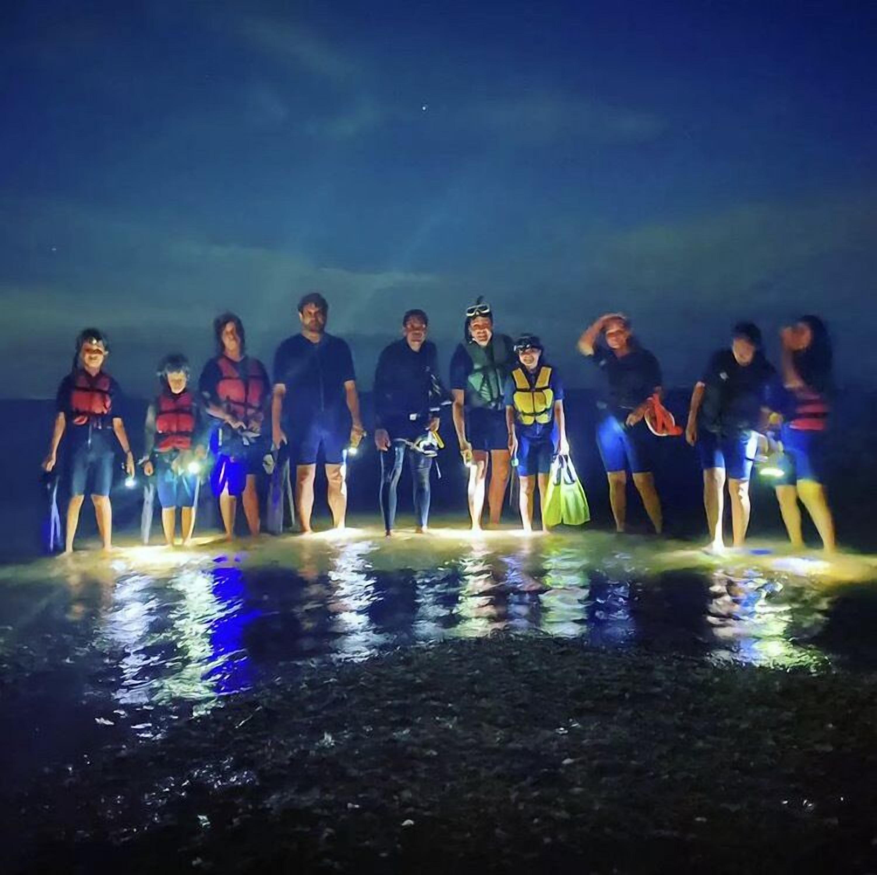 Un groupe de personnes se tient sur une plage la nuit, et la lumière qu'elles émettent se reflète sur le sable mouillé.
