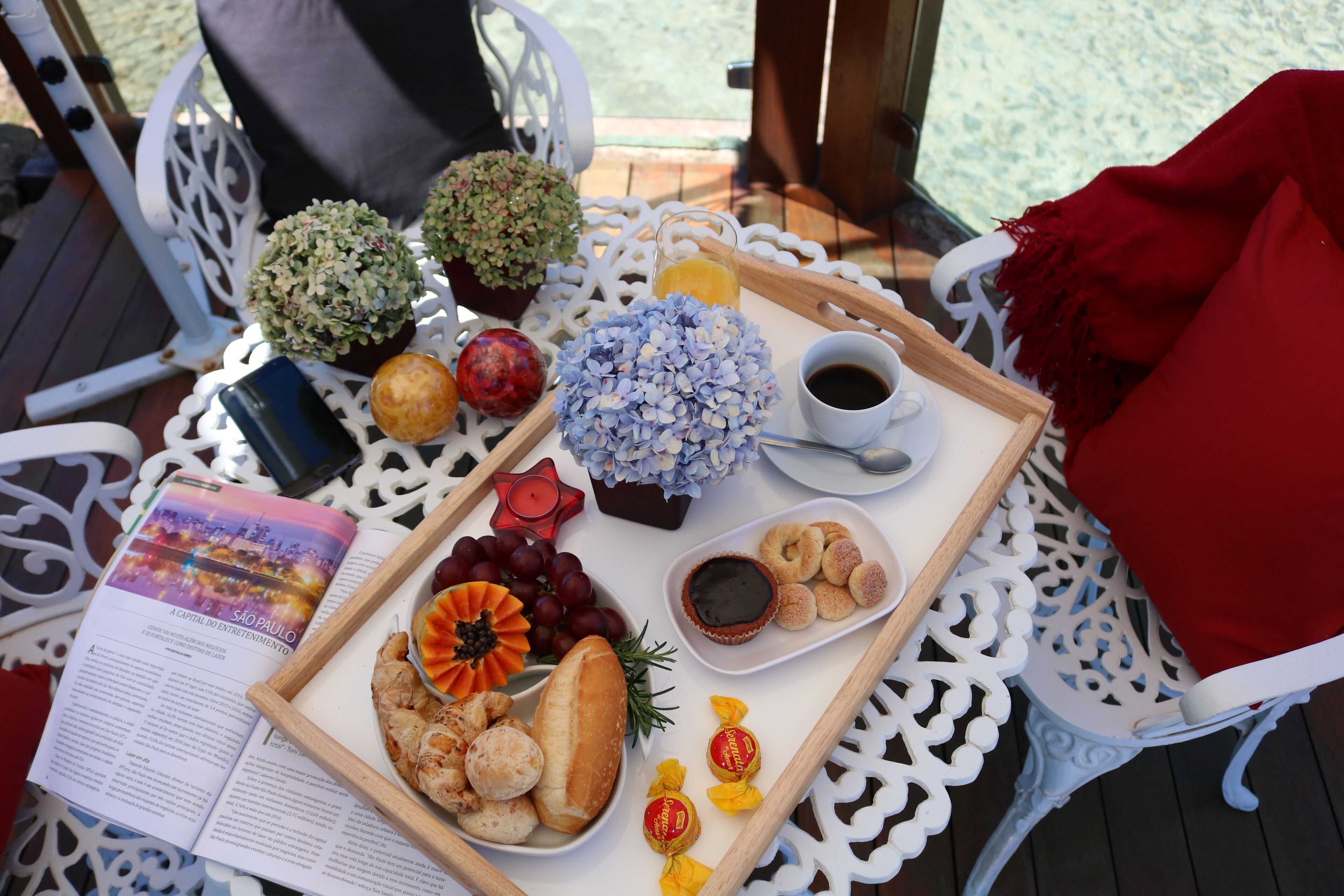 A table with a tray of food, including bread, grapes, and a small dish of jam, set on a white outdoor table with a book and decorative items.