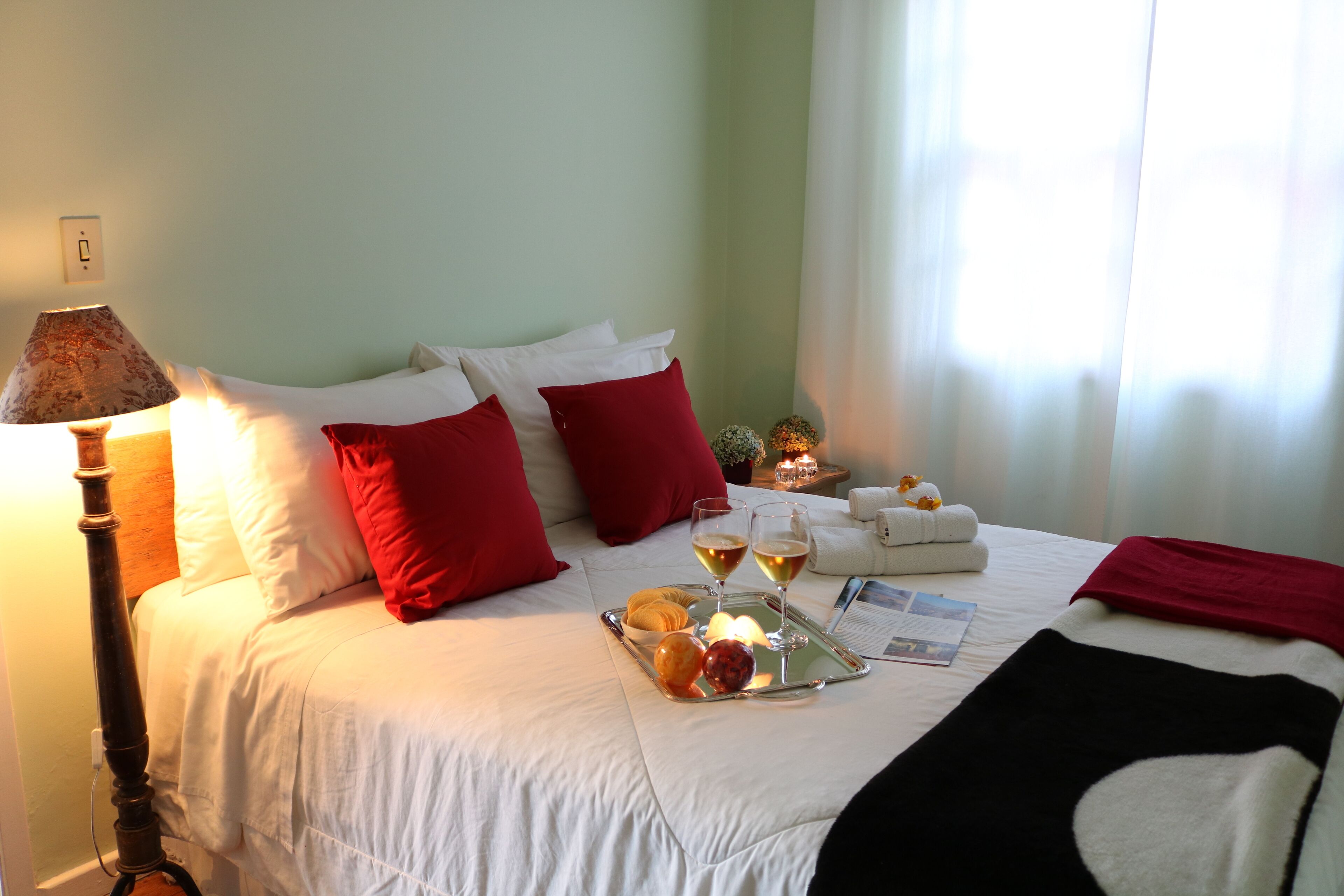 A neatly made bed with white and red pillows, a tray with fruits and drinks, and a lamp beside it.