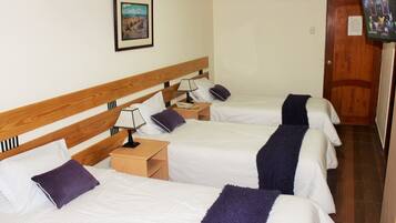 Habitación de hotel con tres camas individuales, cabeceros de madera, ropa de cama blanca, almohadas moradas, mesitas de noche con lámparas, un cuadro enmarcado en la pared y un televisor montado.
