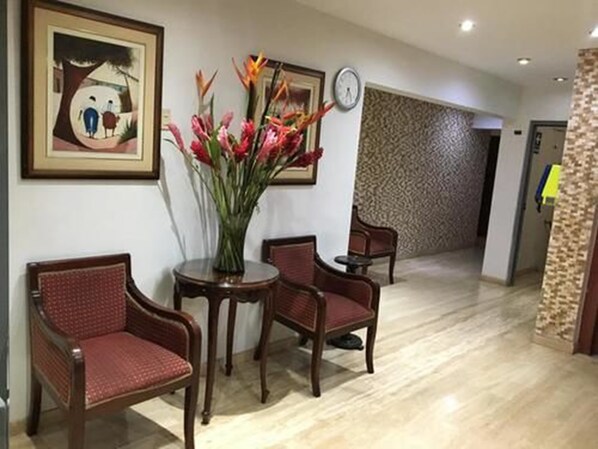Un hall de hotel con una recepción, dos sillas, un jarrón de flores y un reloj en la pared.