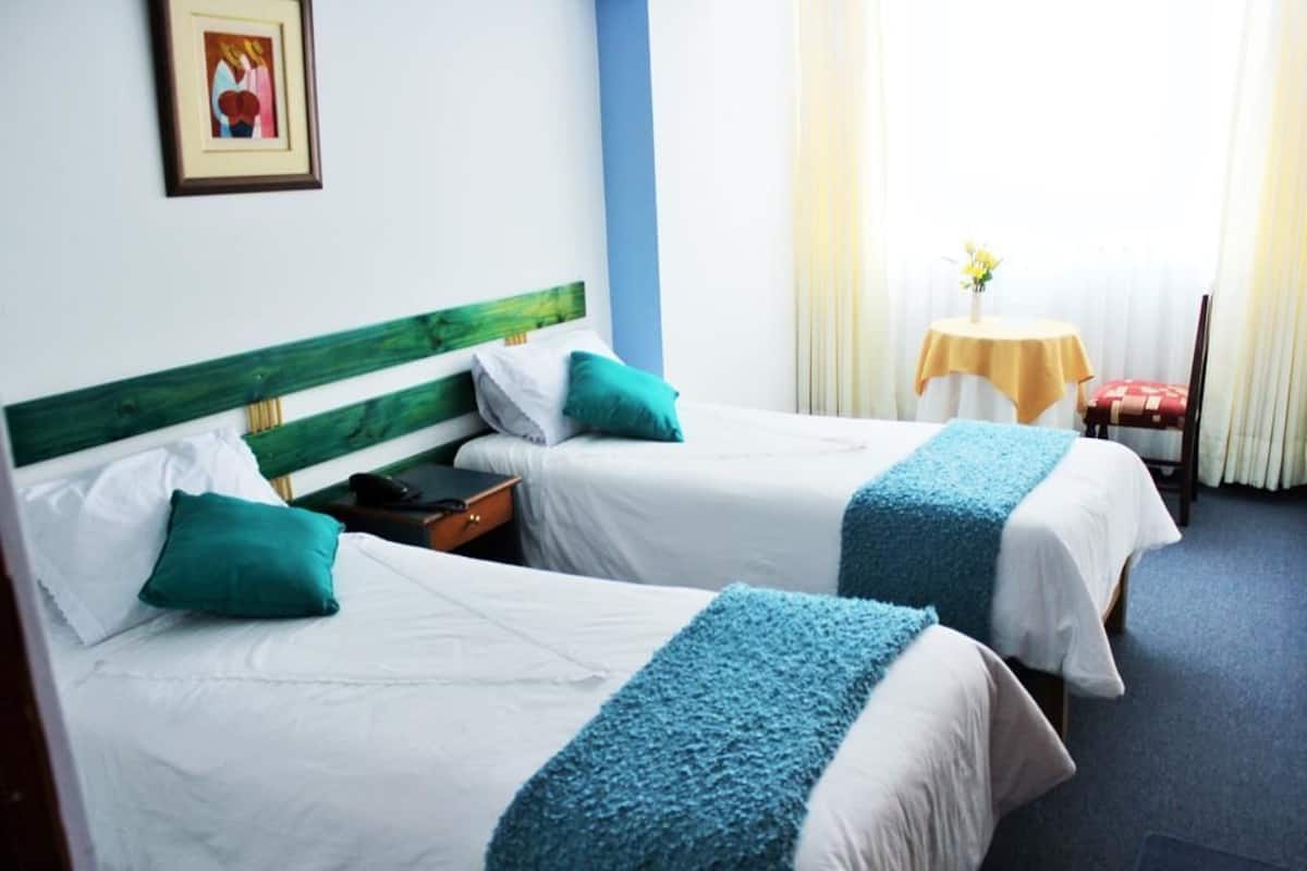 Ein Hotelzimmer mit zwei Betten, jeweils mit einem grünen Kopfteil und weißer Bettwäsche. Ein kleiner Tisch mit gelber Tischdecke und eine Blumenvase sind ebenfalls vorhanden.