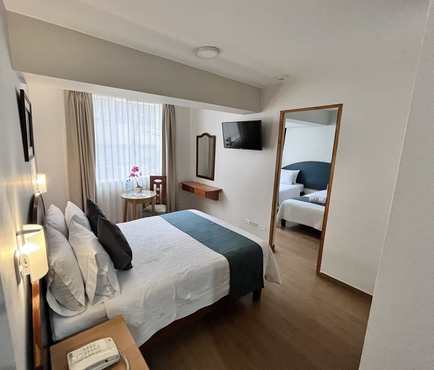 Habitación de hotel con cama, mesita de noche, lámpara, espejo, televisión y una pequeña mesa con una silla.