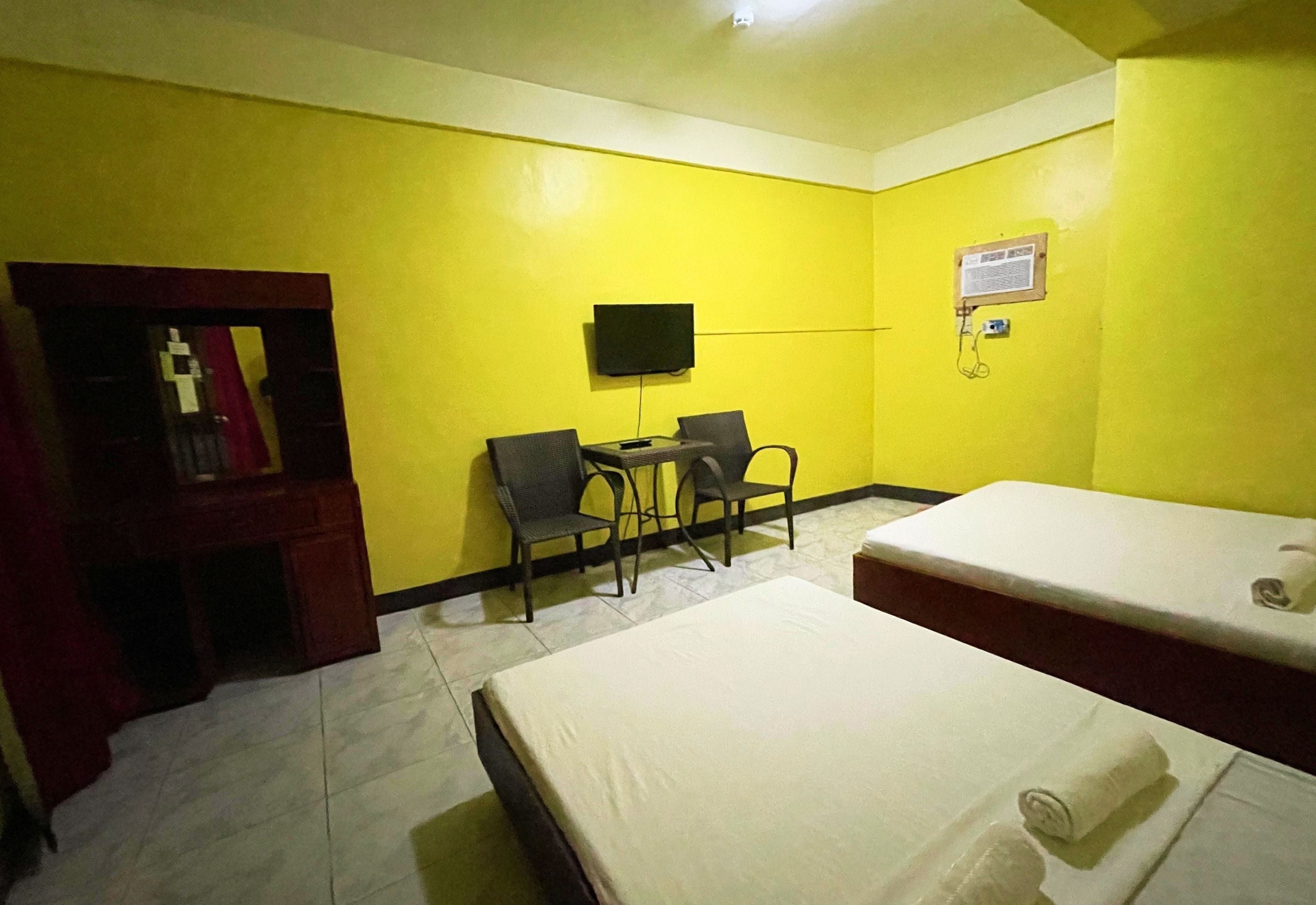 A hotel room with yellow walls, a bed, a desk with two chairs, a television, and a small cabinet.