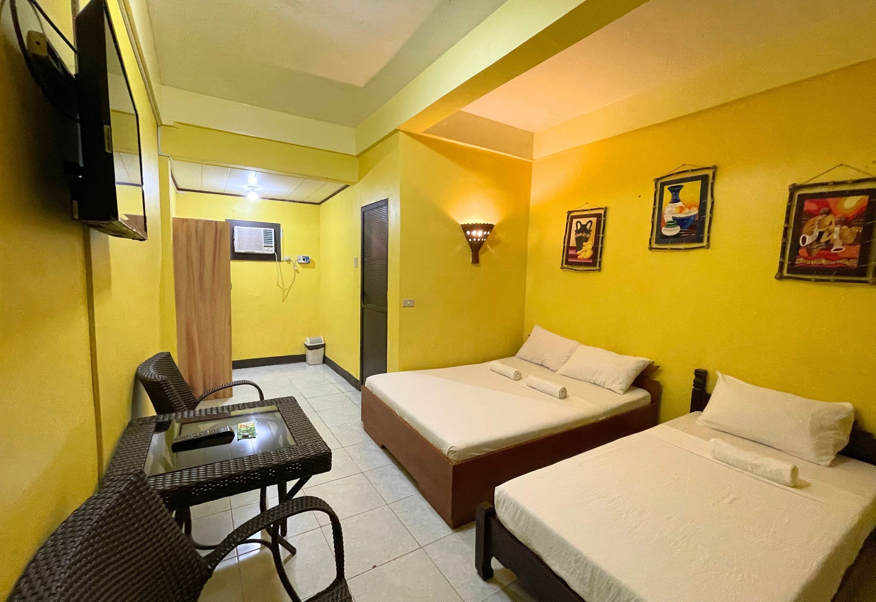 A hotel room with two beds, a small table with chairs, a TV mounted on the wall, and a ceiling fan.