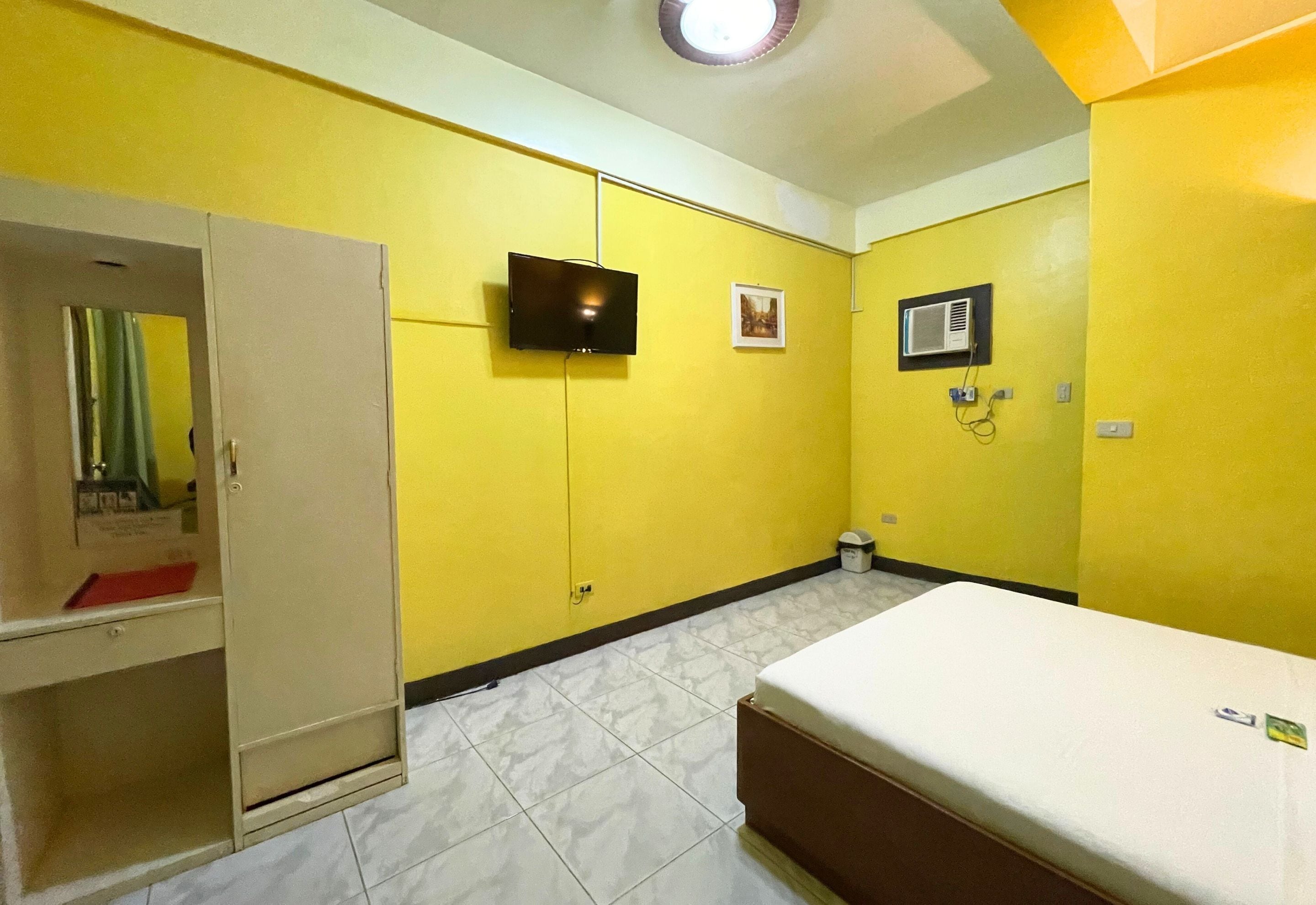 A room with yellow walls, a bed, a wardrobe, a television, and an air conditioning unit.