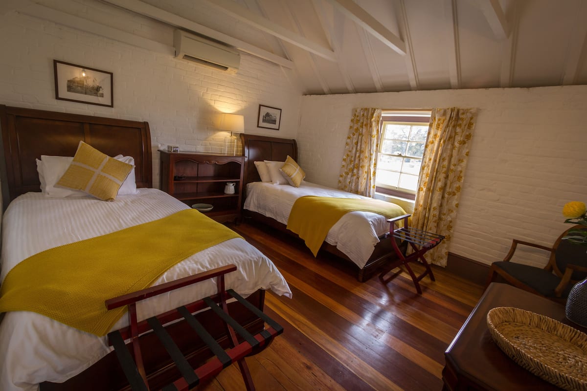 Una habitación con dos camas, suelo de madera, una mesita y una ventana con cortinas.