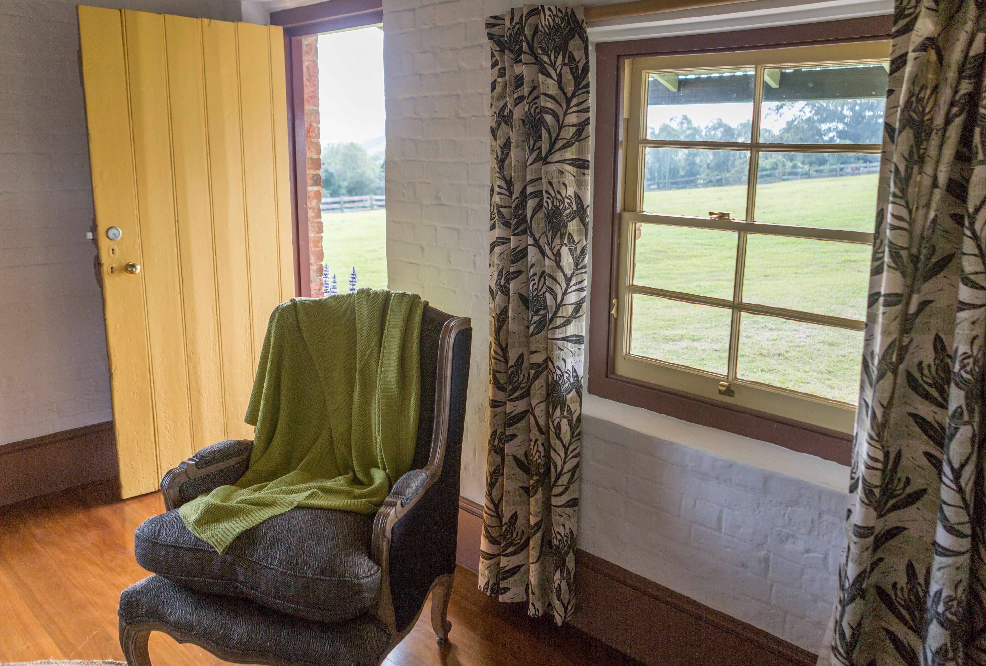 Una habitación con puerta de madera, ventana con cortinas florales y una silla con una manta verde.