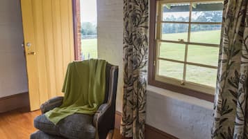 Una habitación con puerta de madera, ventana con cortinas florales y una silla con una manta verde.