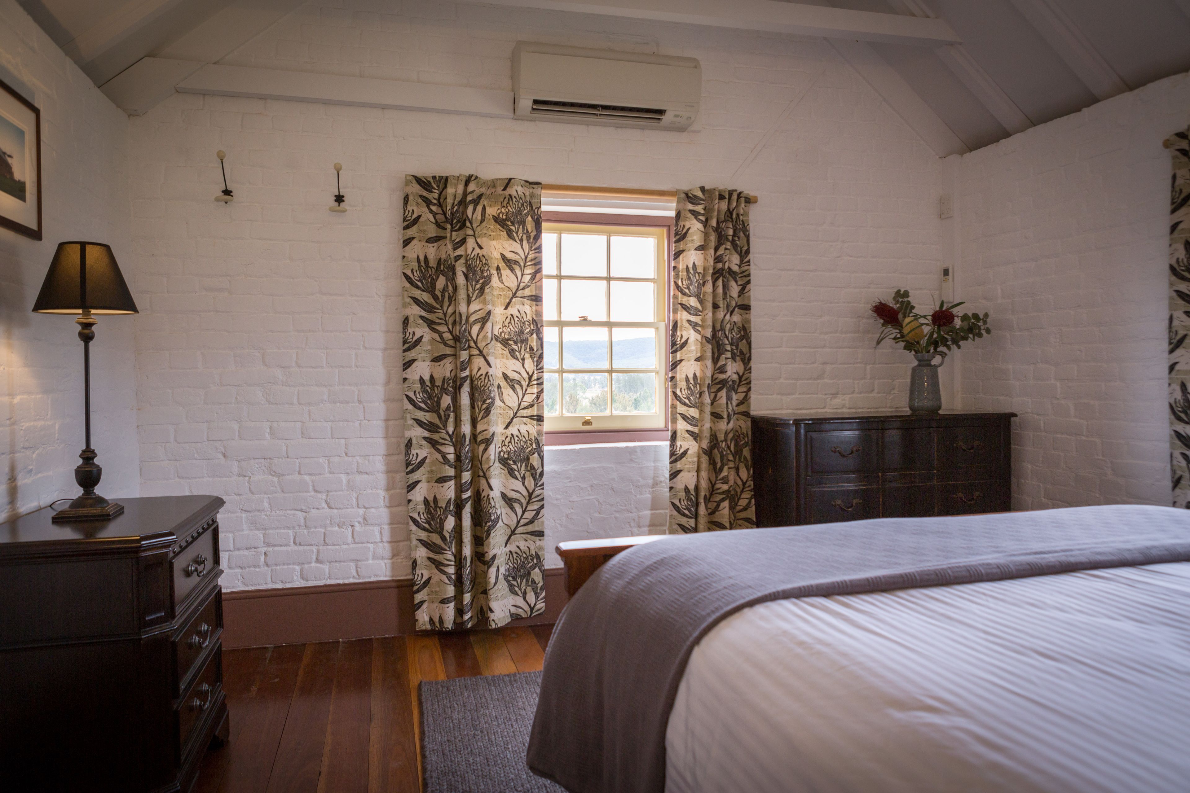 A bedroom with a large bed, a dresser, a lamp, a window with curtains, and a vase with flowers on a dresser.