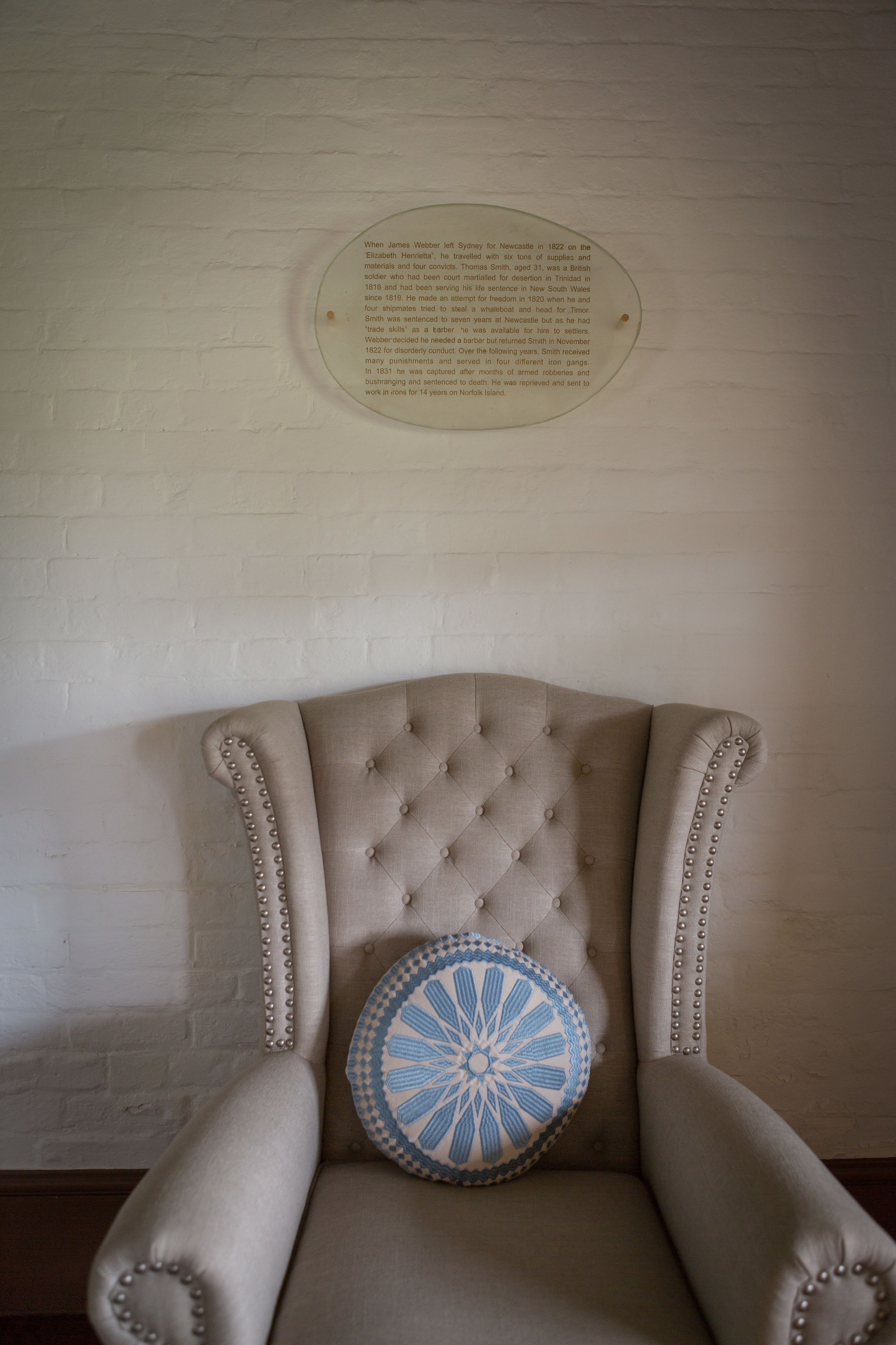 Una silla con respaldo capitonado y almohadones decorativos, ubicada frente a una pared de ladrillo blanco con una placa en la parte superior.