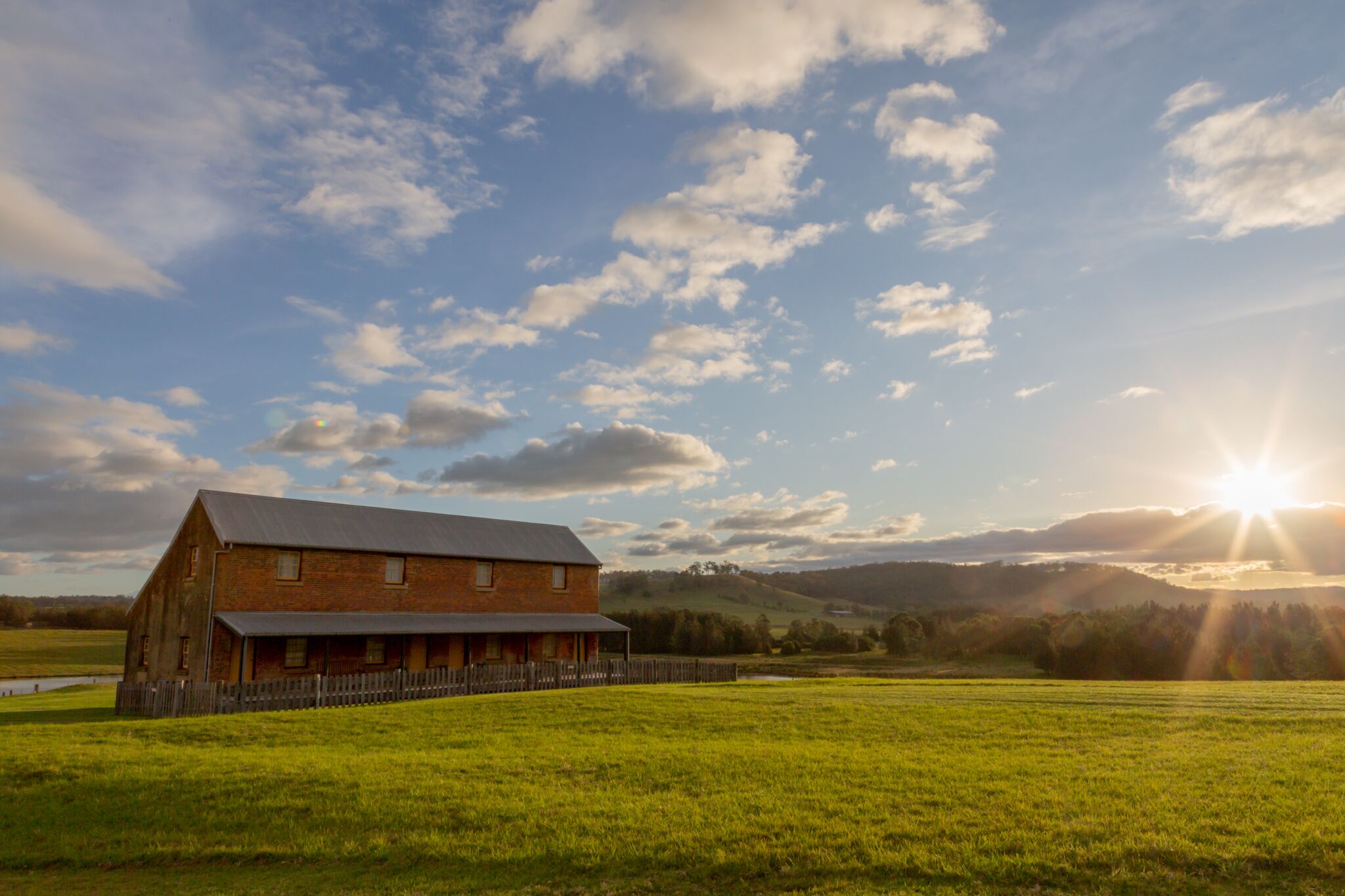 A large brick barn with a fenced area in front, set in a green field with a clear sky and sun.