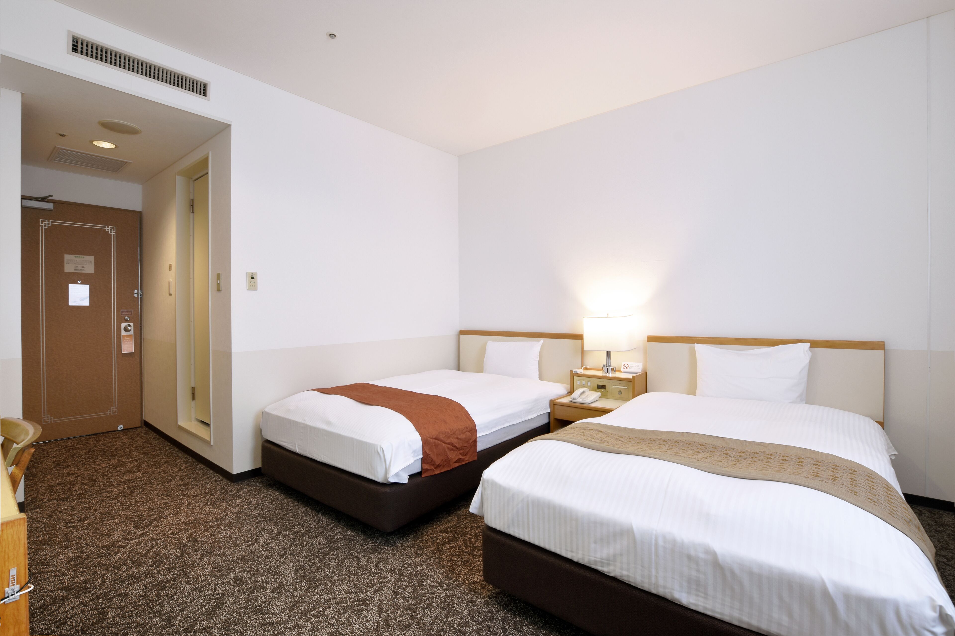 A hotel room with two beds, a desk, a chair, a lamp, and a door.