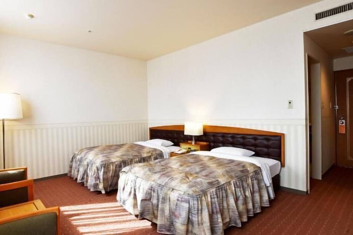 A hotel room with two beds, a desk, a chair, a lamp, and a door.