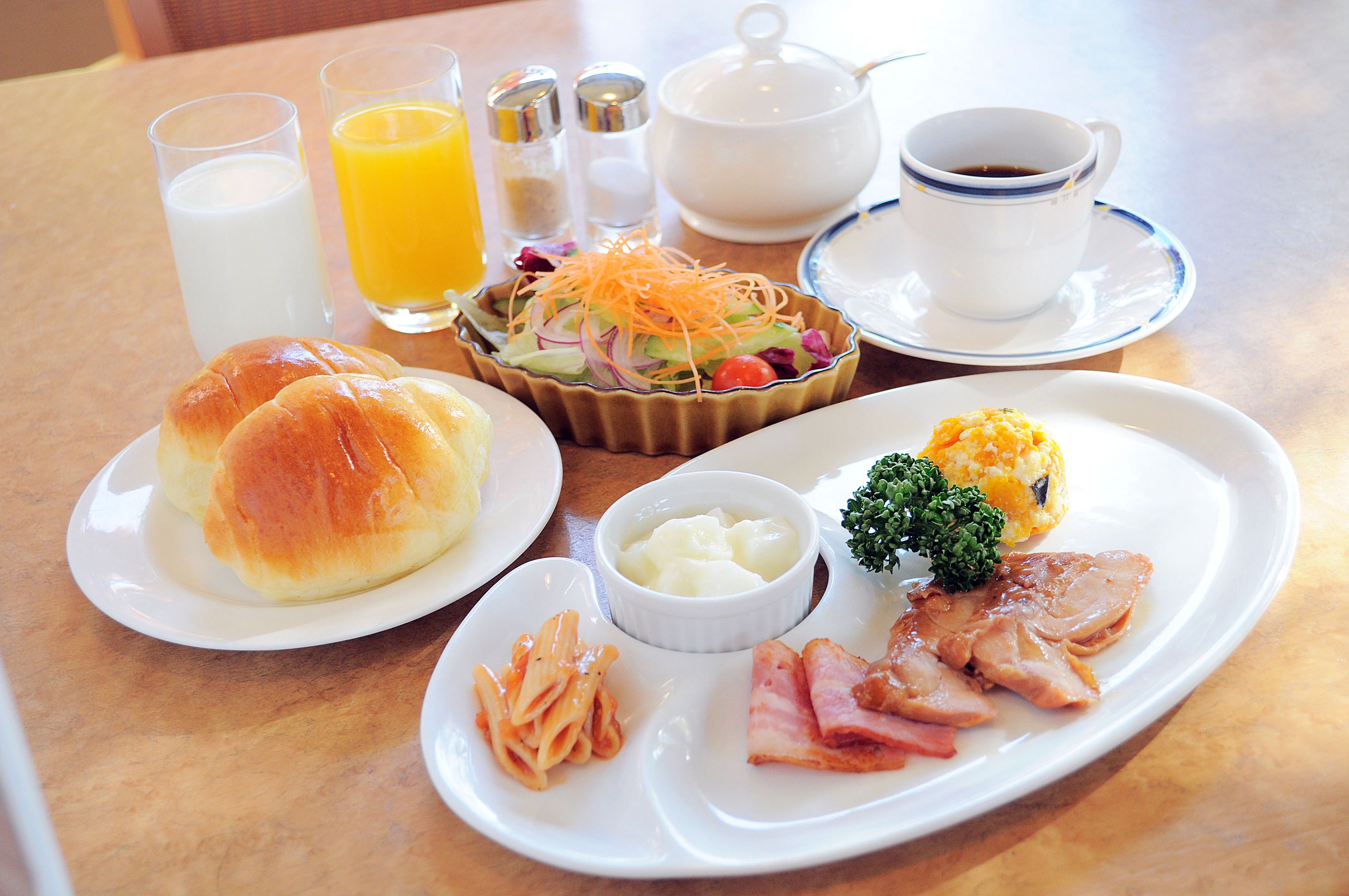 A breakfast table with croissants, ham, pasta, a salad, butter, coffee, milk, and orange juice.