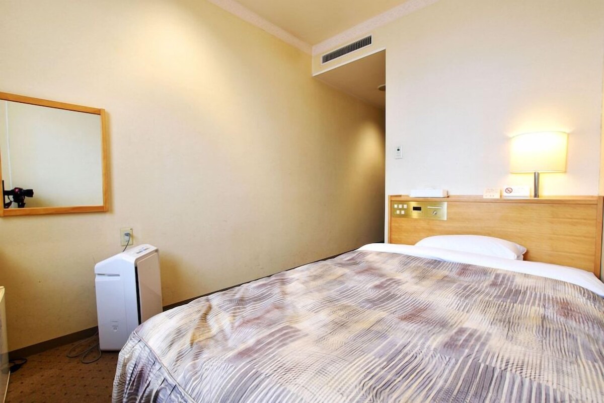 A hotel room with a bed, a bedside table, a lamp, a mirror, and a small white appliance.