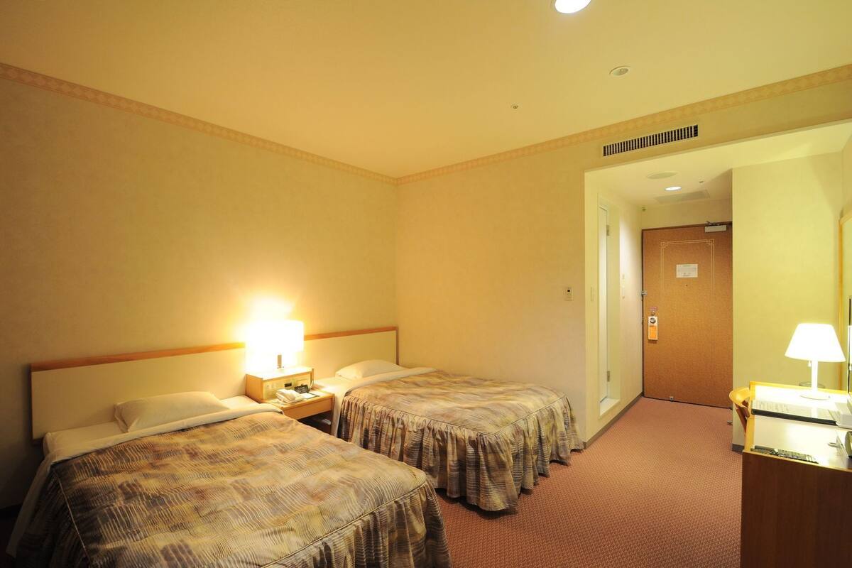 A hotel room with two beds, a desk with a lamp, and a door leading to another room.