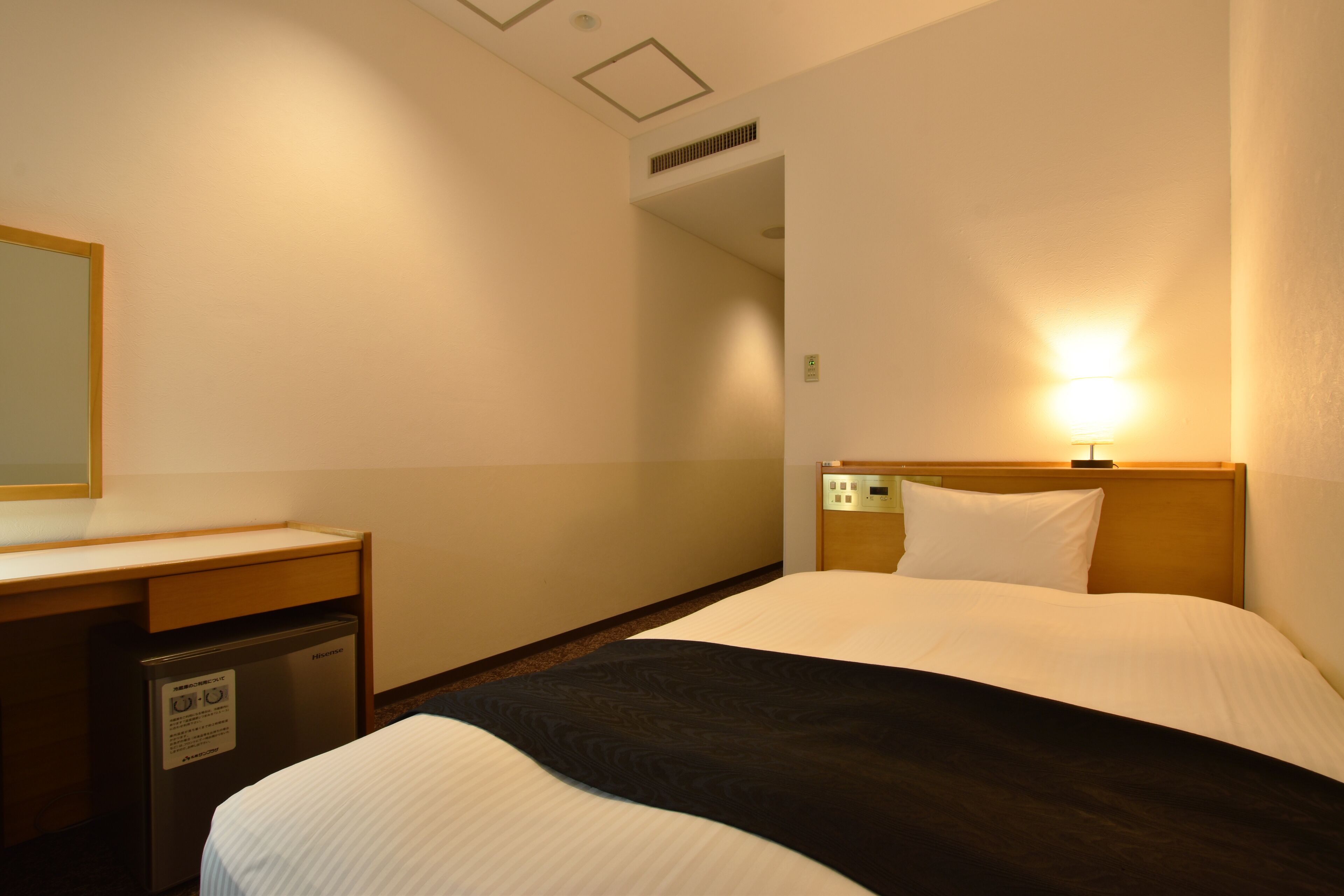 A hotel room with a bed, a desk, a refrigerator, and a nightstand.