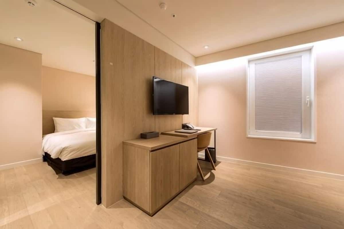 A bedroom with a bed, a TV mounted on the wall, a desk, and a window with blinds.