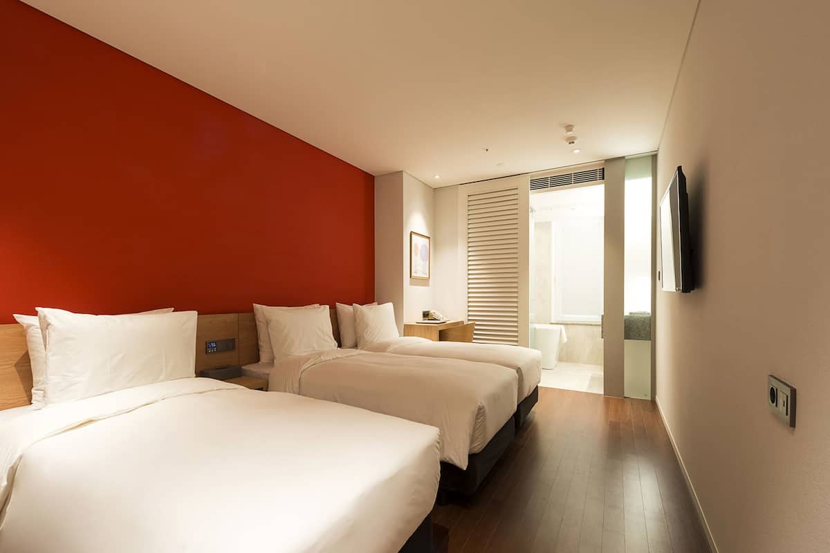 A hotel room with two beds, a red accent wall, a television, and a bathroom visible through a door.