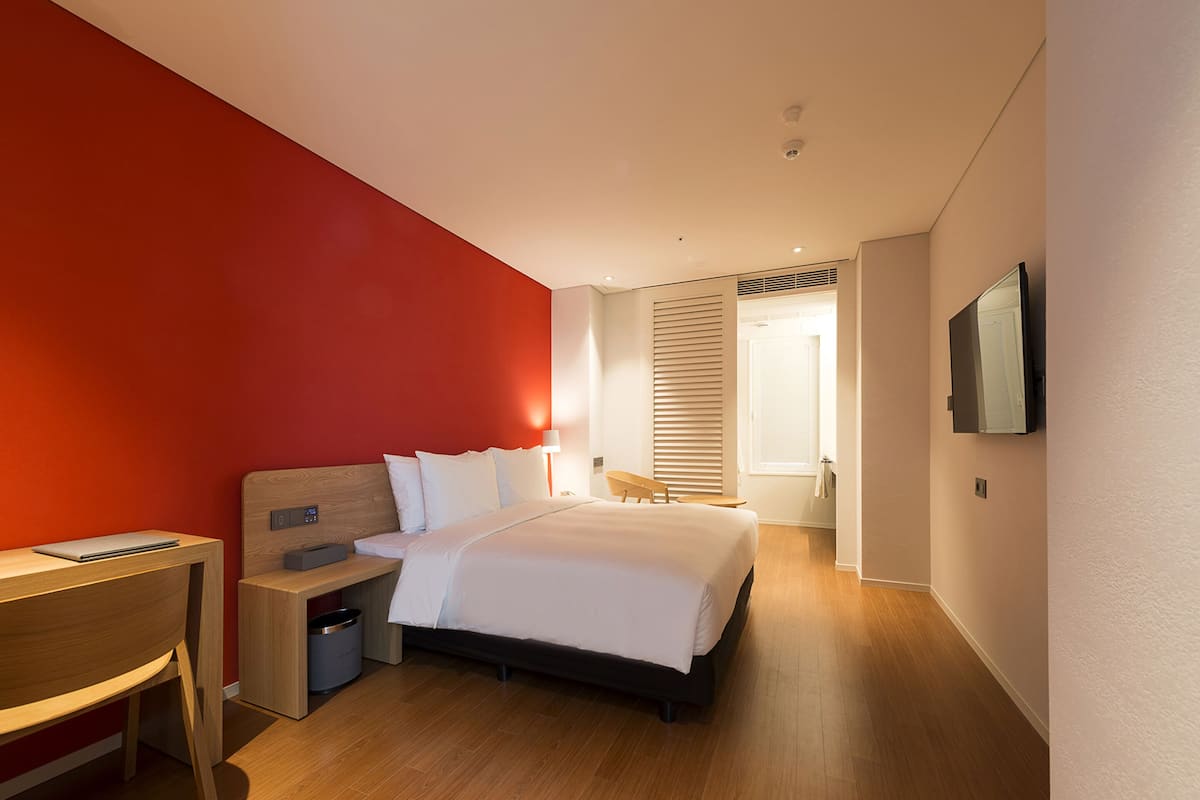 A modern hotel room with a large bed, a desk, a chair, and a TV mounted on the wall.