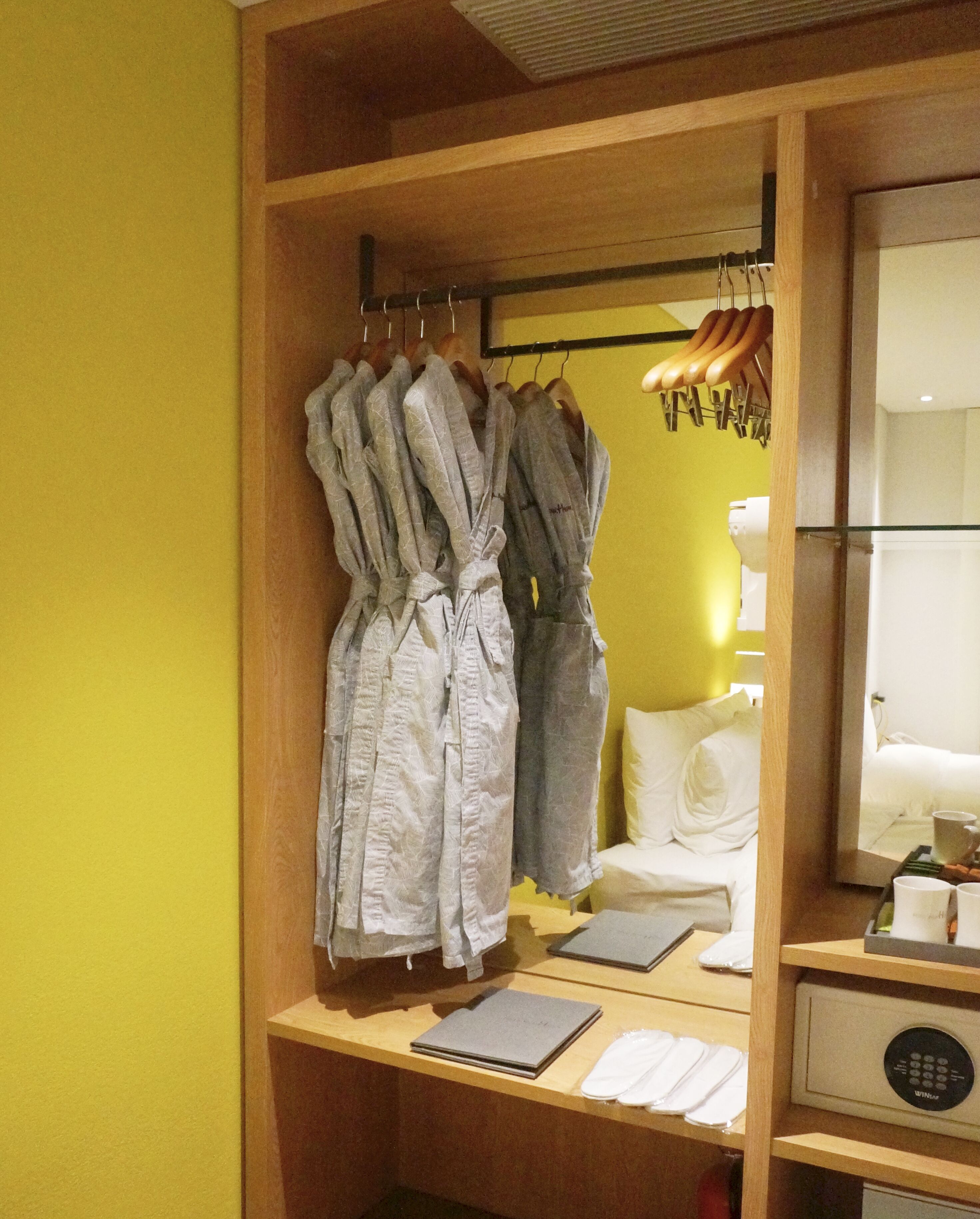 Habitación de hotel con un armario de madera que contiene albornoces, una cama con ropa blanca, una mesita de noche con un cuaderno, una bandeja con tazas y un panel de control.