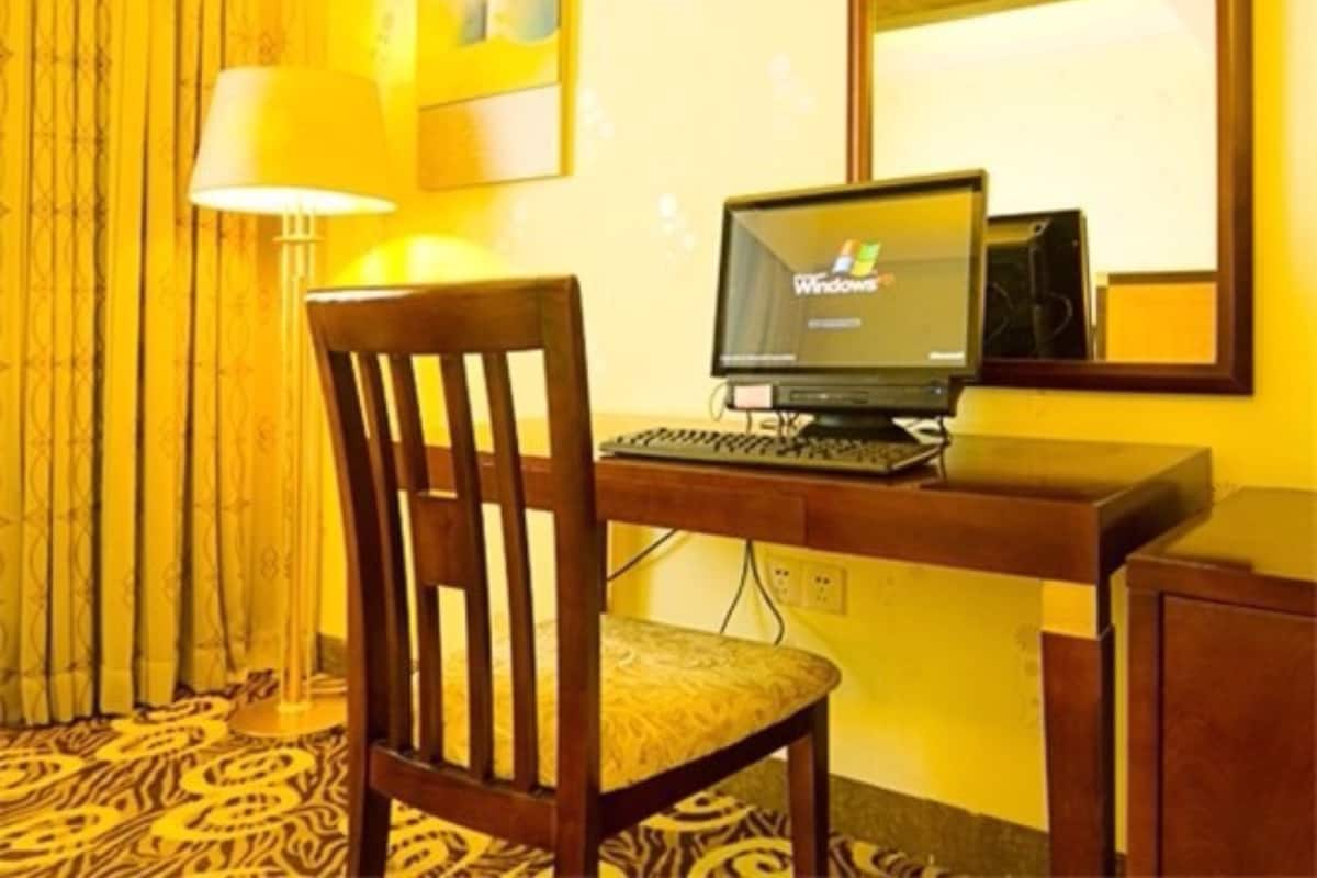 A hotel room with a wooden desk, a computer, a chair, a lamp, and a patterned carpet.