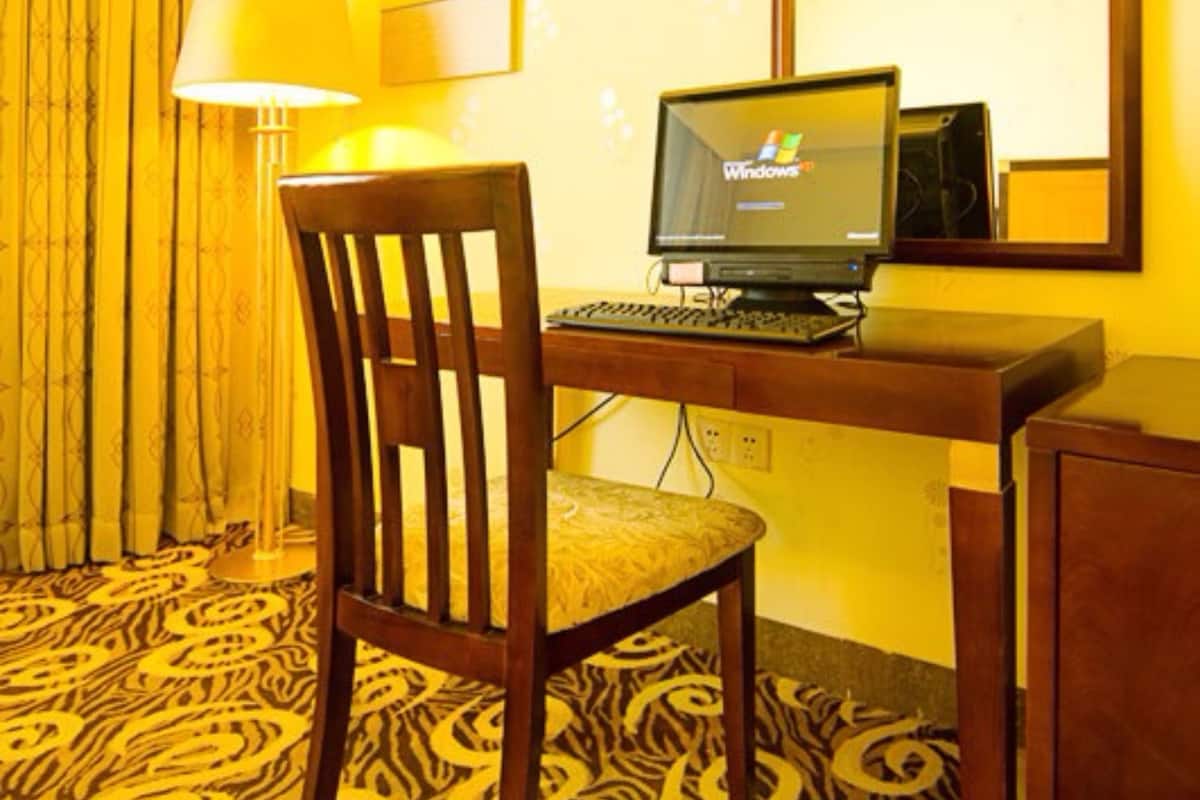 A hotel room with a wooden desk, a computer, a chair, a lamp, a mirror, and a patterned carpet.