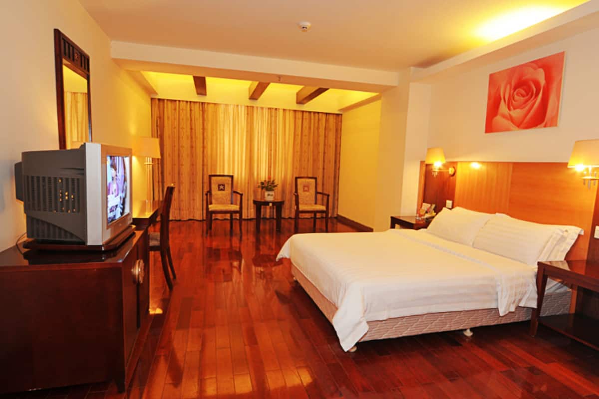 A hotel room with a large bed, a television on a wooden cabinet, two chairs, a small table with a plant, and a painting of a rose on the wall.