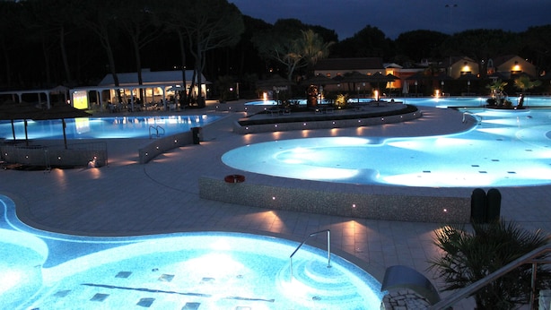 A large outdoor swimming pool area with multiple pools and a central water feature, surrounded by stone walkways and palm trees.