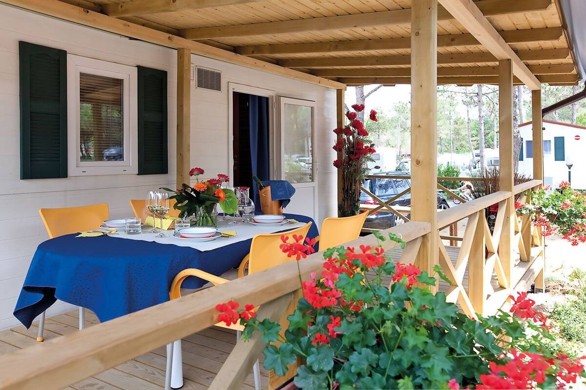 Een overdekte eethoek buiten, met een tafel gedekt voor twee personen, omgeven door houten leuningen en plantenpotten.