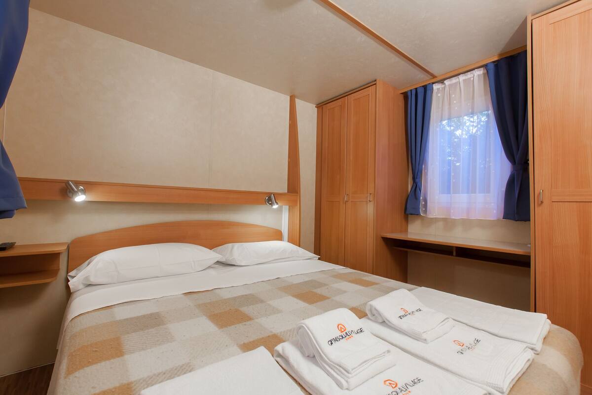Een net opgemaakt bed met een geruit beddegoed, een houten hoofdbord en een raam met blauwe gordijnen.