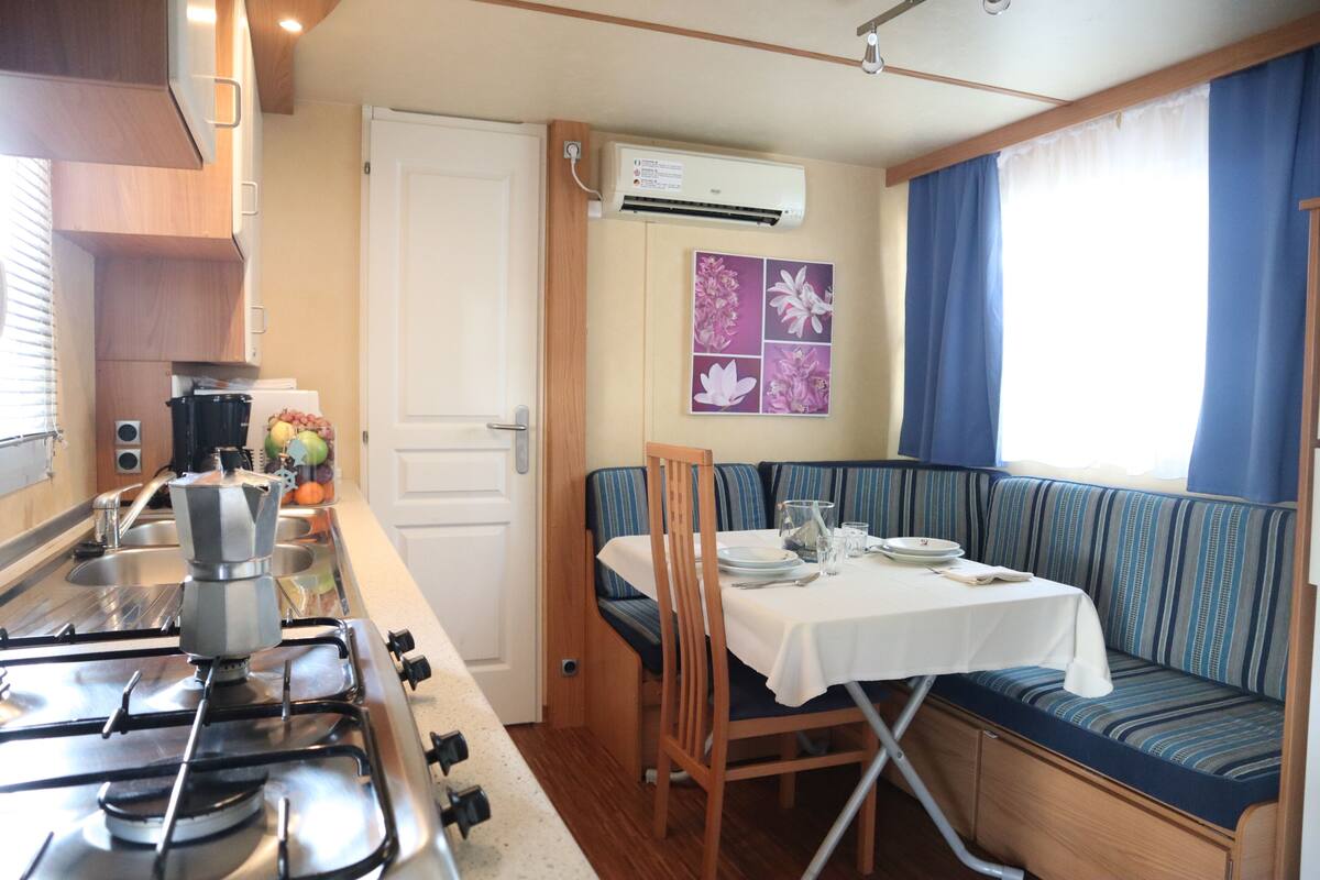 Een compacte kitchenette met een gasfornuis, een spoelbak en een klein aanrecht. Er is een zithoek met een tafel en stoelen, en een raam met blauwe gordijnen.