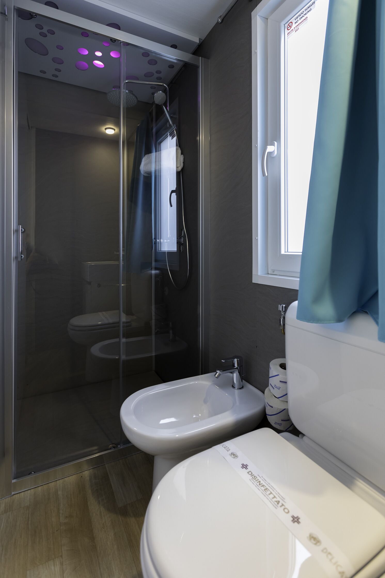 A modern bathroom with a glass shower enclosure, a bidet, a toilet, and a window with a curtain.