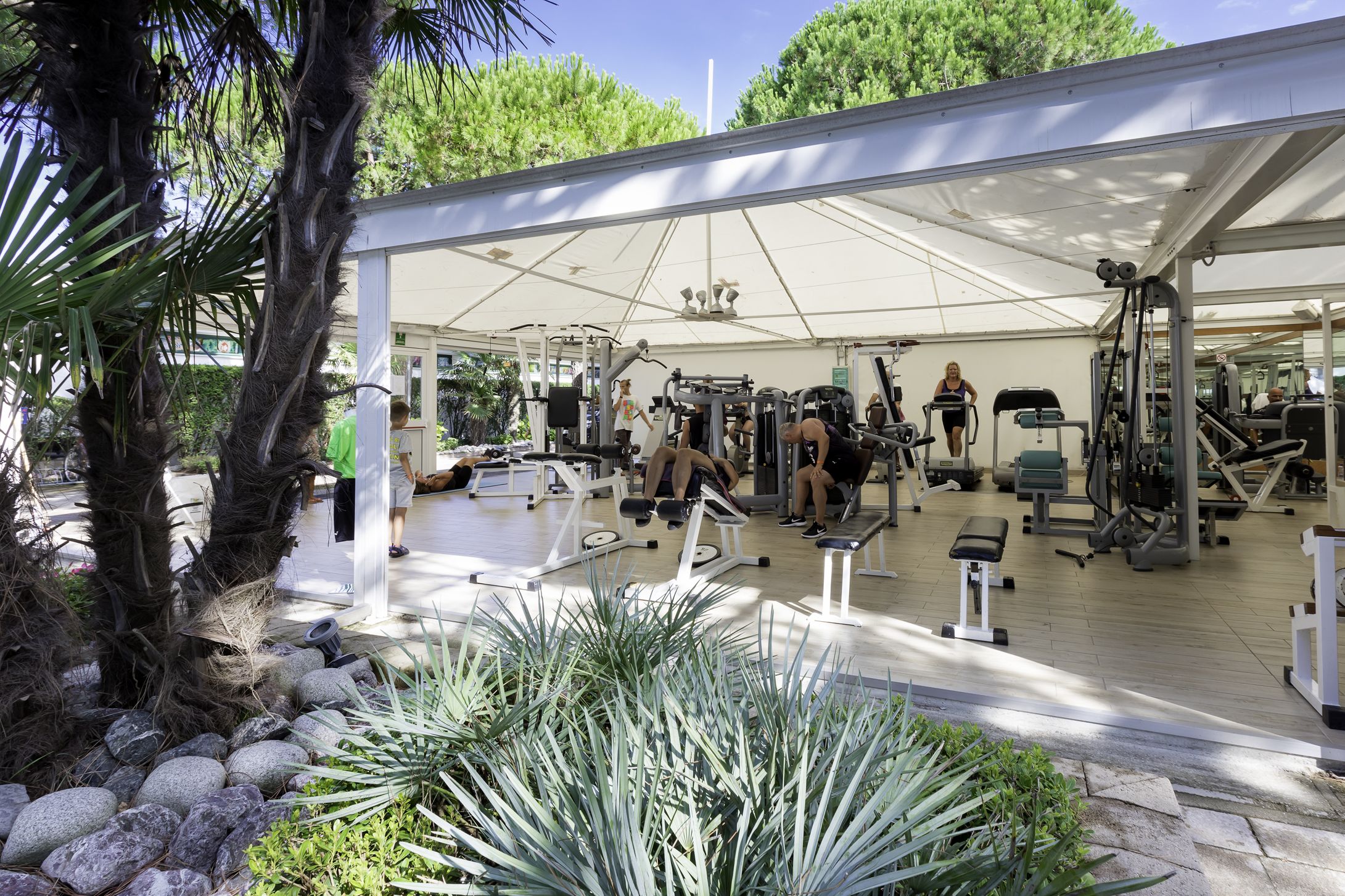 Ein Outdoor-Fitnessbereich mit unterschiedlichen Trainingsgeräten unter einer weißen Markise, umgeben von Palmen und Pflanzen.